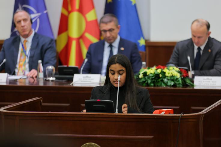 Young woman speaks at a conference table with officials and flags in the background.