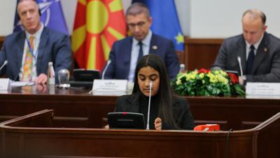 Young woman speaks at a conference table with officials and flags in the background.