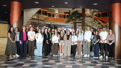 Group of people standing indoors with a staircase and plants in the background.