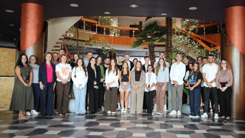 Group of people standing indoors with a staircase and plants in the background.