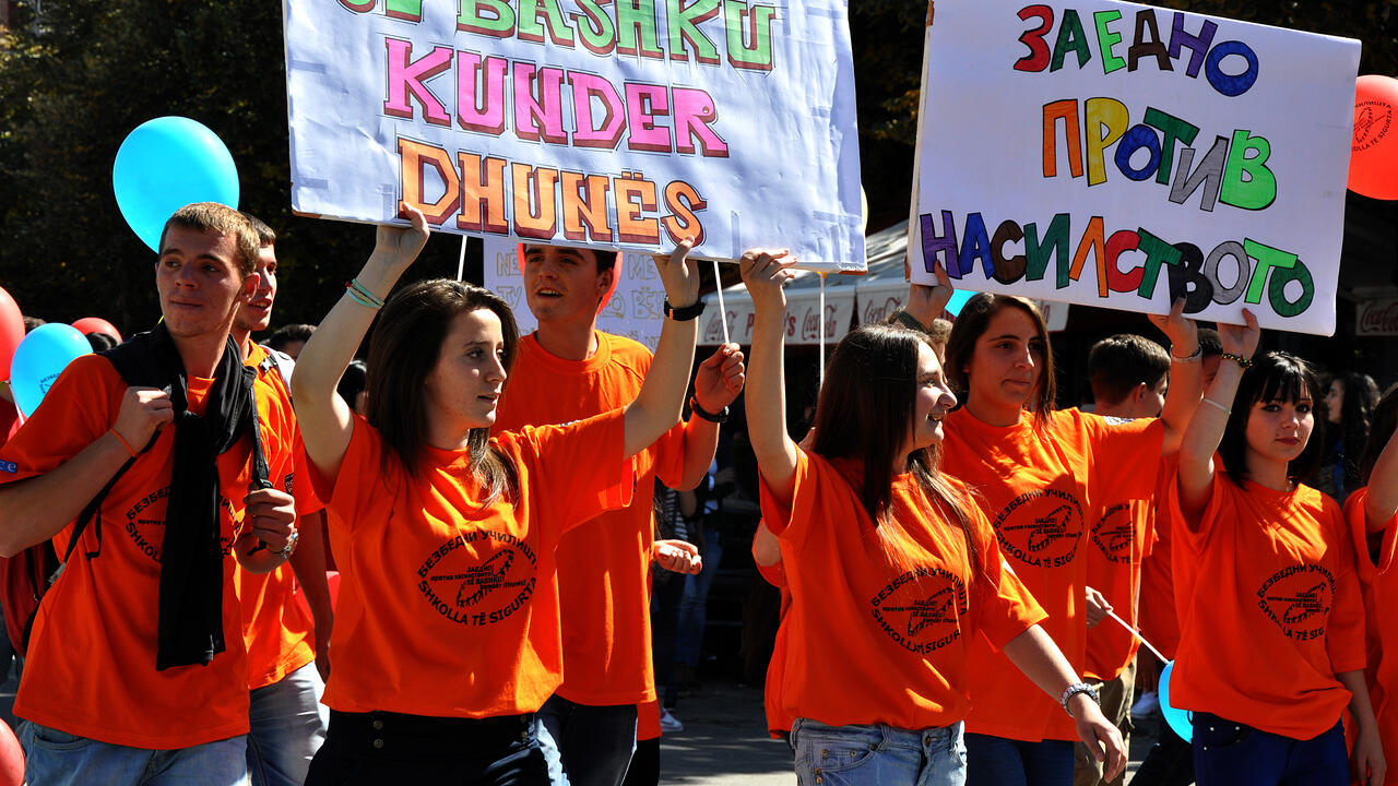 People in orange shirts holding protest signs, marching with colored balloons on a sunny day.